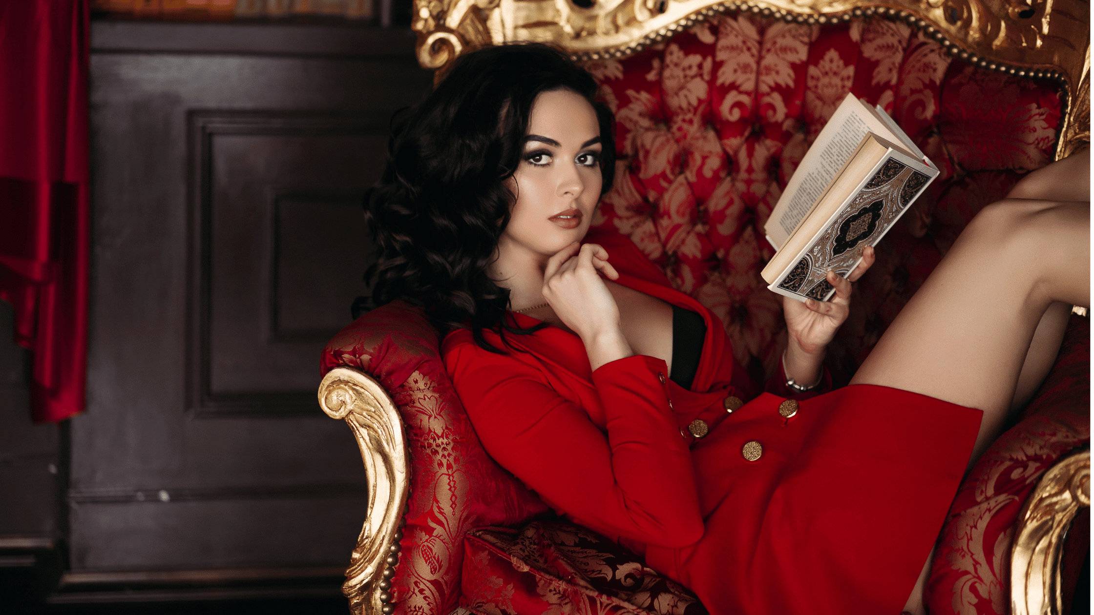 Portrait of girl in short red dress lying on arm chair, holding book in hand